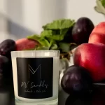 A clear glass container candle with plums and rhubarb in the background